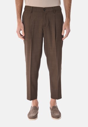 Uomo che indossa pantaloni marroni su misura e mocassini in camoscio marrone, in piedi davanti a uno sfondo bianco neutro.