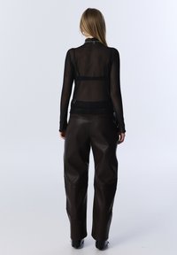 Schwarzes, transparentes Langarmoberteil mit hohem Kragen, kombiniert mit dunkelbraunen, strukturierten, weitgeschnittenen Hosen. Bemerkenswerte Details umfassen den enganliegenden Schnitt.