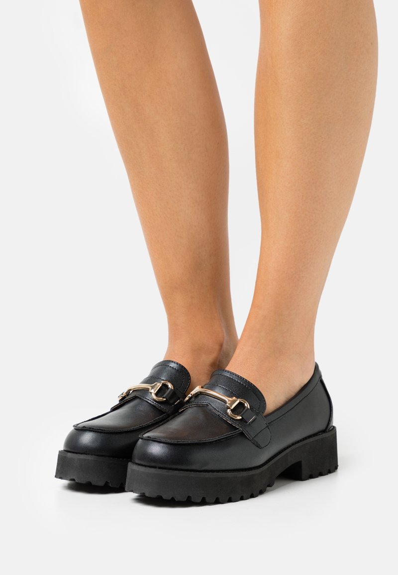 Mocassins en cuir noir avec une semelle épaisse, dotés d'une boucle dorée et d'un bout arrondi. Texture lisse avec un design à enfiler.