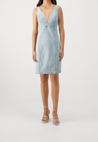 Valkoinen vaaleansininen denimi mekko, jossa on syvä V-kaulus, olkaimettomat hihat, tiukka siluetti ja hienovarainen saumaus, yhdistettynä hihnatossuihin vaaleassa beigestä.