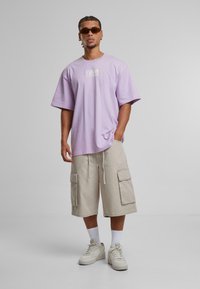 Light purple oversized t-shirt with a logo, beige cargo shorts with side pockets, and white sneakers. Simple, relaxed design with subtle texture.