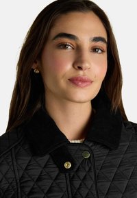 Sieviete ar gariem brūniem matiem valkā zelta auskarus un melnu kviltētu jaku ar korduroja apkakli, izskatās nedaudz smaidīga, raugās uz priekšu.