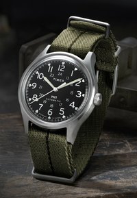 Timex MK1 automatisch polshorloge met zwarte wijzerplaat, witte cijfers, lichtgevende wijzers en groene stoffen band op een houten oppervlak.