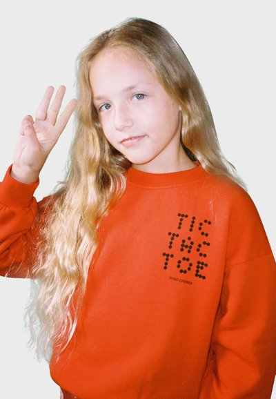 Rode sweatshirt met pofmouwen, met een zwart tic-tac-toe ontwerp op de voorkant. Lang, golvend blond haar en lichtblauwe ogen.