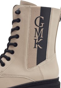 Bottine en cuir synthétique beige avec accents noirs, dotée d'œillets noirs, d'un panneau latéral texturé et d'un logo "GMK".