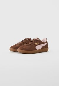 Bruna mockaskinnsneakers från Puma med rosa sidostreifen och gummisulor, placerade sida vid sida på en vit bakgrund.