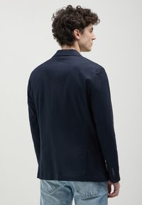 Blazer navy in tessuto liscio, con spacco singolo, rever a punta e dettagli ai polsini con quattro bottoni. Indossato sopra jeans azzurri.