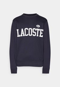 Lacoste marinblå sporttröja med vit "LACOSTE"-text. Rund halsringning, ribbad mudd och nederkant. Har en liten broderad logotyp på bröstet.