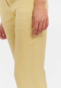 Pantalon jaune clair à la texture lisse, avec une coupe ajustée et une couture sur le côté, accompagné d'un délicat bracelet en or porté à la main.