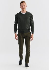 Ciemnozielony sweter w serek, lekki dzianinowy, w połączeniu z oliwkowymi spodniami. Model w czarnych sneakersach, stojący na tle jednolitego białego tła.