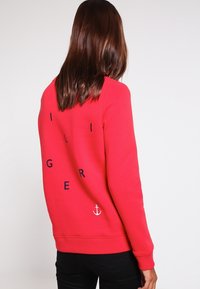 Červená mikina s dlouhými rukávy, s nápisem v tmavě modré barvě a malým bílým kotvou v dolní části. Bavlněná směsová tkanina.