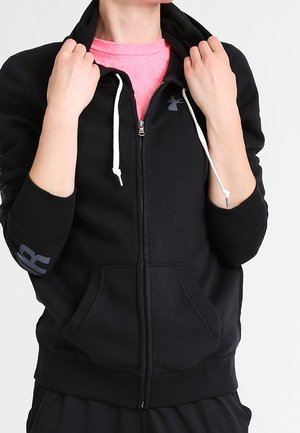 Persona vistiendo una sudadera negra con cremallera y cordones blancos sobre una camiseta rosa, sosteniendo la capucha cerca del cuello.