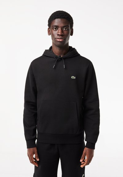 Hoodie noir en tissu doux avec une poche kangourou devant, un capuchon à cordon de serrage réglable, et un logo vert sur la poitrine gauche.