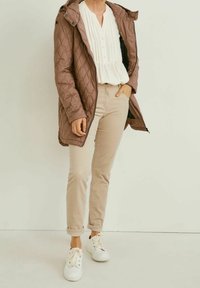 Person iført en quilted brun jakke over en hvid bluse, beige bukser med opsmøg og hvide sneakers, der står foran en ensfarvet baggrund.