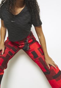 Kvinna i svart Nike-tröja och röda mönstrade byxor med en liten svart väska fäst, lutar sig framåt mot en vit bakgrund.