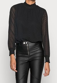 Schwarze Bluse mit transparenten, gemusterten langen Ärmeln, gerafftem Ausschnitt und strukturierten Manschetten, kombiniert mit schwarzen, hoch geschnittenen Lederhosen.