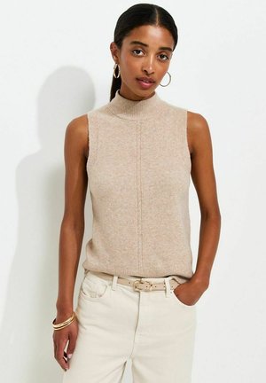 Donna con capelli scuri in un top beige senza maniche di maglia e pantaloni chiari, con orecchini a cerchio e braccialetti d'oro, che posa con una mano in tasca.