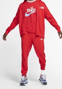 Osoba ubrana w czerwony dres Nike z bluzą na zamek i spodnie dresowe, w połączeniu z wielokolorowymi sportowymi butami i białymi skarpetkami.