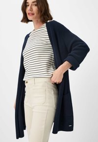 Navy knitted cardigan with open front, ribbed sleeves, and pockets, worn over a white and navy striped top and light beige pants.