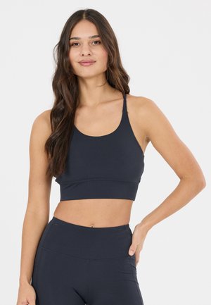 Ung kvinde med langt mørkt hår iført en sort sports-bh og leggings med høj talje, stående med den ene hånd på hoften.