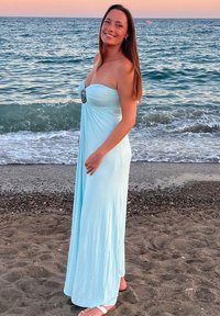 Himmelblaisses, trägerloses Maxikleid mit gerafftem Oberteil und dekorativem silbernen Akzent. Getragen mit weißen Sandalen, stehend am Strand.