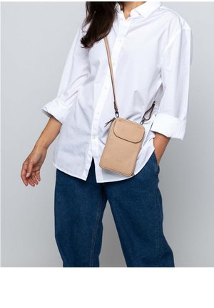 Frau trägt ein weißes Hemd mit Knöpfen und blaue Jeans mit einer beigen Umhängetasche, die an ihrer Hüfte hängt.