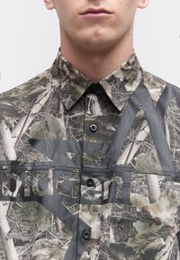 Mann trägt ein camouflagegemustertes Hemd mit Knöpfen, versehen mit Blatt- und Astmotiven sowie teils sichtbarem, kräftigem schwarzem Text auf dem Stoff.