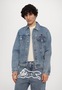 Giacca di jeans di un blu chiaro con un taglio classico, presenta due tasche sul petto e un logo rosso applicato. Abbinata a jeans di jeans blu chiaro con una grafica bianca.