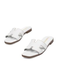 Sandalias deslizantes blancas con recortes en forma de H, suela plana, acento marrón y detalle de costura beige a lo largo de los bordes. Textura de material suave.
