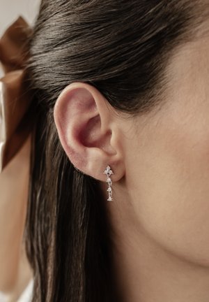 Silver drop earrings featuring a floral design with small clear gems, hanging below the lobe, set against dark, straight hair.