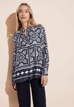 Camicia a righe navy con colletto e maniche lunghe, caratterizzata da motivi floreali e geometrici bianchi. Abbinata a pantaloni in denim scuro.