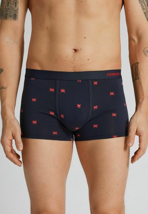 Mężczyzna w granatowych bokserkach z czerwonymi wzorami krabów i widocznymi tatuażami na ramionach, stojący na jednolitym tle.