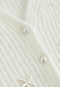 Bílý pletený cardigan s žebrovanou strukturou, perlami na knoflíky a dekoračním akcentem. Měkký materiál s útulným vzhledem.