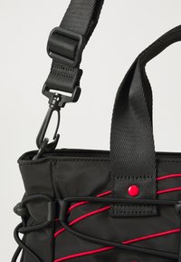 Sac en tissu noir avec une surface texturée, présentant des accents en cordon élastique rouge, une bandoulière robuste et une fermeture à bouton-pression.