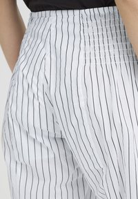 Gros plan sur la taille et les hanches vêtues d'un pantalon blanc à fines rayures verticales noires avec une ceinture élastique froncée.