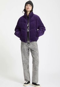 Doudoune en velours côtelé violet avec un col montant, fermeture éclair et motif matelassé, portée sur un t-shirt graphique et associée à un jean gris.