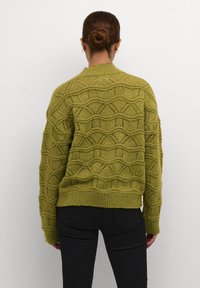 Gestrickter grüner Pullover mit Wellenmuster, ausgestattet mit gerippten Bündchen und Saum sowie einer groben Textur. Von hinten gesehen, getragen zu dunklen Jeans.
