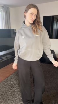 Siva majica s kapuljačom s prednjim džepom i logotipom, uparena s crnim širokim hlačama. Tkanina s teksturom, opušteni kroj i ležerni stil.