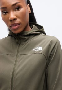 Φούτερ μπουφάν με κουκούλα σε ελιά πράσινο χρώμα με απαλή υφή, με λευκό λογότυπο στο στήθος και πλήρες φερμουάρ. Κοντινή εικόνα του προσώπου ενός μοντέλου.