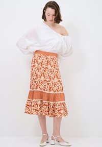 Ein orange- und cremefarbenes, gemustertes Kleid mit Blumenmuster, Rüschensaum und karierten Akzenten, kombiniert mit einem weißen, schulterfreien Oberteil und High Heels.