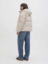 Beige pufferjakke med hætte, der har en kasseformet design, kviltet tekstur og lange ærmer, båret over lyseblå brede jeans.