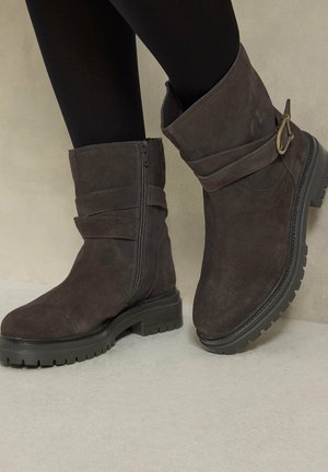 Bottines en daim marron foncé avec fermeture éclair latérale, boucle décorative, semelle épaisse à crampons, portées avec des collants noirs sur fond clair.