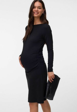 Femme enceinte en robe noire cintrée tenant une pochette noire, souriante, debout devant un fond clair uni.