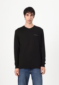 T-shirt noir à manches longues en coton, avec un col rond, une coupe ajustée, et un petit logo blanc sur la poitrine gauche.
