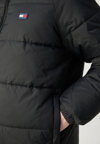Doudoune noire avec surpiqûres horizontales et logo Tommy Jeans sur la poitrine ; la main de la personne est dans la poche latérale.