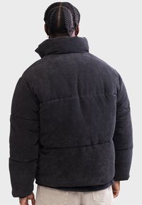Chaqueta de plumas de pana negra con un alto cuello, que presenta secciones acolchadas horizontales y una textura acogedora, vista desde la parte posterior.