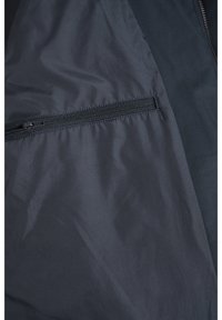 Tamsiai mėlynas šiurkštus švarkas, turintis užtrauktukų kišenę, švelnią audinio tekstūrą ir minimalistinį dizainą su kontrastingomis plokštėmis.