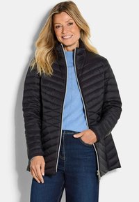 Vrouw die glimlacht, in een zwart gewatteerd jack over een lichtblauwe sweater en donkerblauwe jeans, met één hand in de zak tegen een witte achtergrond.