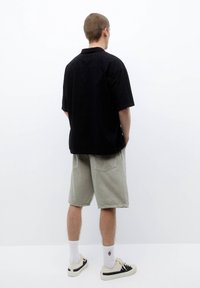 Maglietta nera a maniche corte con design testurizzato, abbinata a pantaloni corti verde chiaro. Le scarpe sono beige con dettagli neri e i calzini bianchi sono visibili.