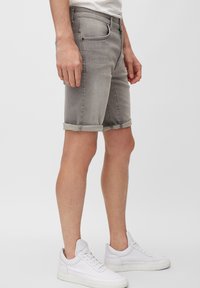 Pantalones cortos de mezclilla gris con puños enrollados, diseño de cinco bolsillos y frente plano. Llevados con zapatillas blancas sobre un fondo sencillo.